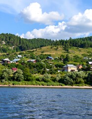 Fototapeta premium A picturesque riverside village nestled on a hillside, with colorful houses and lush greenery, beneath a vibrant sky filled with fluffy clouds.