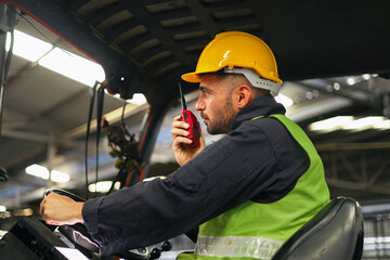 man in  yellow helmet and green vest is talking on  walkie talkie. He is wearing  hard hat and is driving  forklift