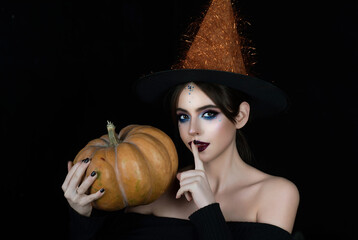 Witch posing with pumpkin in black studio. Young woman in witch hat celebrating Halloween. Sexy...