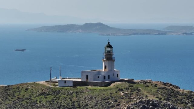 Drone orbit around Mykonos Lighthouse capturing stunning views of Delos island and Chora village under a bright sunny sky, showcasing the beauty of the Aegean Sea and Cyclades.