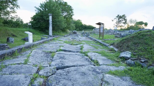 Slow motion push in shot along a Roman stone road with grass between the stones at Nicopolis ad Istrum, surrounded by greenery and cloudy sky