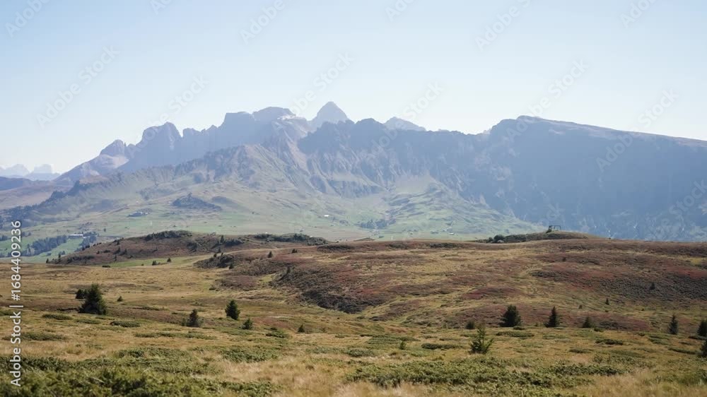 Poster wide view of open grassy field with scattered shrubs and trees beneath jagged mountain range in dolo
