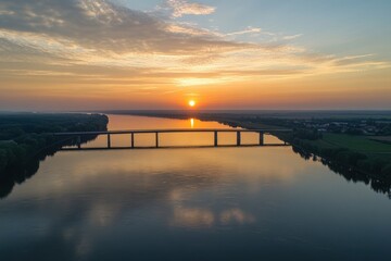 Obraz premium Serene Sunset Over Tranquil River Reflecting Golden Light with a Distant Bridge Silhouette.