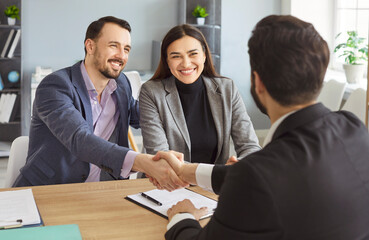 Happy smiling couple shaking hands reaching agreement with a man realtor or financial advisor signing successful contract sitting at desk on workplace. Insurance agent consulting clients in office.