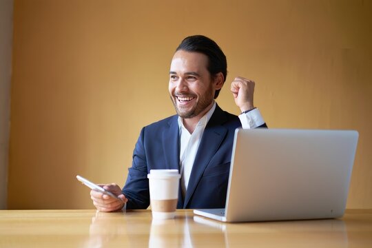 Successful businessman celebrating achievement at desk with laptop and coffee cup near him