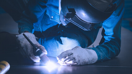 A skilled worker is welding metal joints in an industrial workshop. The helmeted figure is focused on the task, with sparks flying and blue light illuminating the workspace