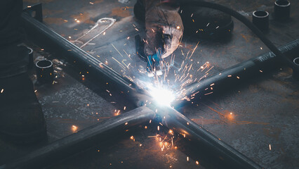 A welder focuses on metalwork in a workshop, using a welding torch. Bright sparks illuminate the...