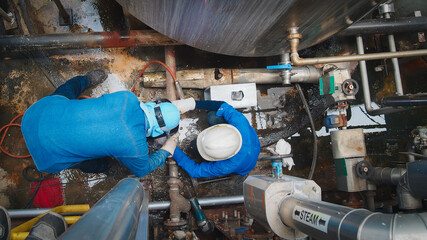 Two workers in protective gear are engaged in maintenance tasks on a complex piping system. They...