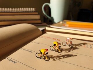 Mini toy of action figure at table with blurred background. Miniature diorama photography concept design. Minifigure of bicycle athlete character at office desk with book and pencil.