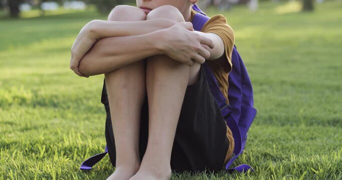 Sad boy sits on green grass in park with his knees hugged to his chest and backpack behind him. Kids thoughtful face shows light tension and reflection. Childhood emotions, self-reflection, psychology
