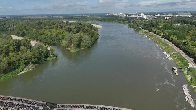 drone pulls away and camera tilting down over iron bridge between cremona - lombardy and castelvetro piacentino . emilia romagna over Po river, with riverbanks lined by greenery.