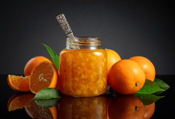 Orange jam in glass jar and fresh fruits on a black reflective background.