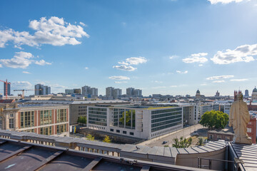 Berlin skyline with modern blocks and historic domes in the distance. A statue overlooks the city under a bright blue sky.