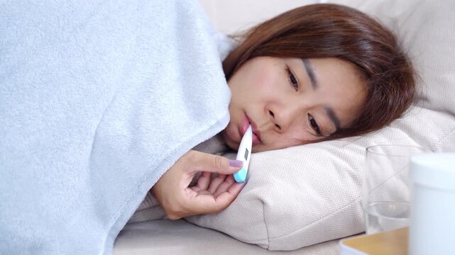 Sick Asian woman lying on sofa at home, wrapped in a blanket, checking thermometer in her mouth, looking weak and exhausted, suffering from high fever fatigue, and flu or cold symptoms, COVID-19