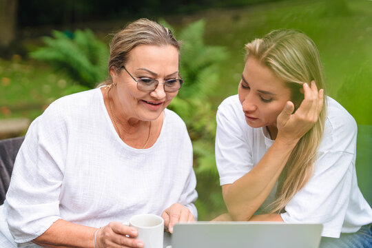 Enjoying a sunny afternoon, two women share moments of joy while exploring a laptop outdoors in a serene garden setting