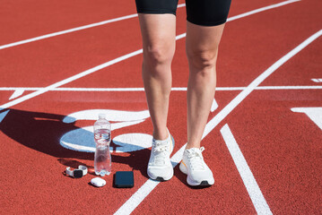 Sportswoman's feet in sneakers, reusable glass water bottle, headphones and smartphone on the red sports carpet. Sports concept of water balance for active training on a sunny day.