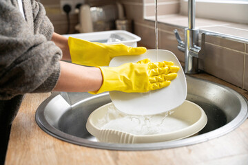 Person does dishes in a home kitchen during daytime, wearing yellow gloves and focused on cleaning plates in a sink filled with soapy water