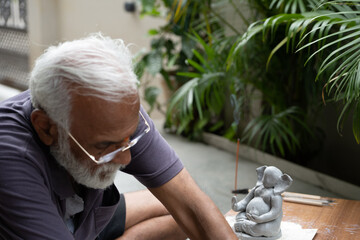 Indian old man crafting an eco-friendly clay Ganesh idol for Ganesh Chaturthi and teaching kids how...