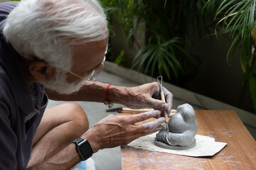 Indian old man crafting an eco-friendly clay Ganesh idol for Ganesh Chaturthi and teaching kids how to do it, sitting outdoors in a garden
