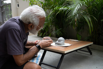Indian old man crafting an eco-friendly clay Ganesh idol for Ganesh Chaturthi and teaching kids how to do it, sitting outdoors in a garden
