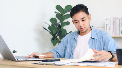Fototapeta premium Young Asian man working on laptop at home office desk with books and plants in background. Concept of remote work, freelance business, and digital lifestyle.