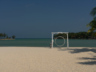 empty sandy beach with a large white circular swing frame near the shore, calm turquoise sea and...