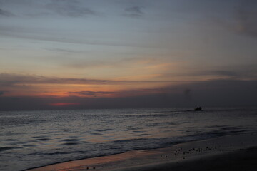 Sunset over calm ocean casting warm glow across the water. Beach sandy under sky is lightly clouded...