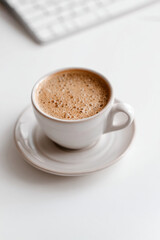 Fototapeta premium White Cup of Frothy Coffee on Saucer with Keyboard in Minimal Workspace