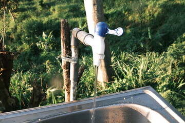 Fresh clean water flowing from a rustic outdoor tap into a metal sink under warm sunlight.