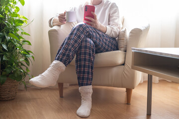 Woman wearing pajamas and wool socks is relaxing on an armchair at home, enjoying a hot beverage...