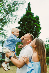 Fototapeta premium A joyful family moment unfolds as a father lifts his cheerful son high in the air, while the loving mother laughs, all set in a picturesque outdoor scene on a warm, sunny summer day