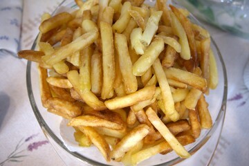 Golden crispy French fries served in a large transparent glass bowl on a table, close-up view.
