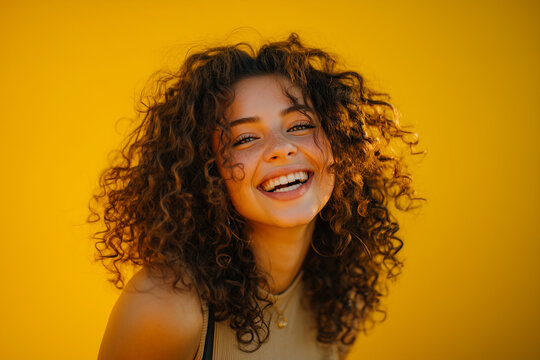 Joyful young woman with curly hair laughing against vibrant yellow background in natural light