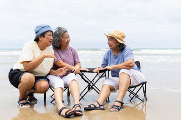 Three elderly women enjoy summer outdoor activities on beach with people sitting on portable chairs...
