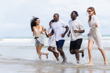 Group of people enjoying summer outdoor activities on beach running and splashing water with happy expressions and casual clothing under bright sky