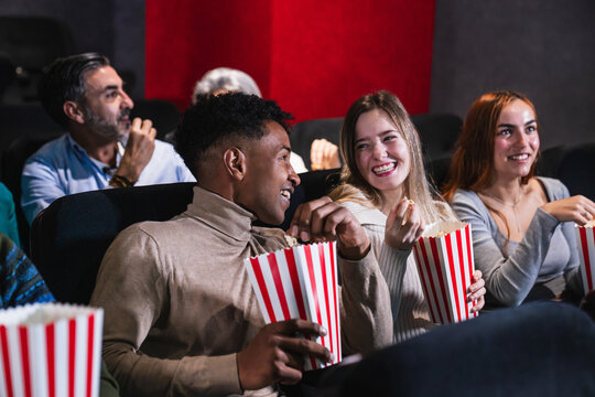 Group of cheerful young friends enjoying a film while sharing popcorn in a cozy movie theater, creating joyful memories together