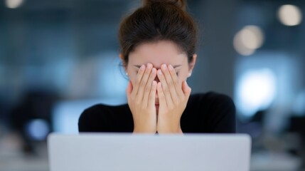 A young woman covers her face in frustration while sitting in front of a laptop. The modern workspace is softly lit, creating a calm yet intense atmosphere. 