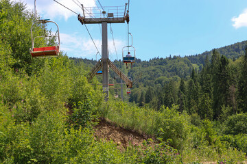 A summer day on a mountain slope, where ski lift chairs move slowly above dense thickets. A person...