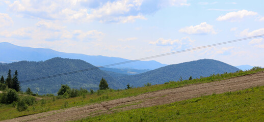 A wide mountain landscape where green, forested hills stretch to the horizon. In the foreground, a dirt patch transitions into grassy slopes, with the sky adorned by light clouds.