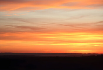 Fototapeta premium A vibrant sunset paints the sky with fiery oranges and soft yellows, casting a warm glow over a silhouetted landscape below.