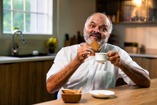 Elderly Indian man dips biscuit in tea at home kitchen