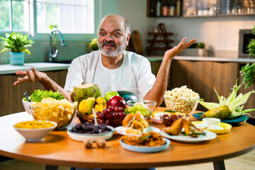 Happy senior Indian foodie enjoying snack-filled table in stylish kitchen setup