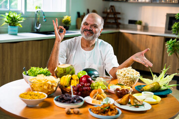 Happy senior Indian foodie enjoying snack-filled table in stylish kitchen setup