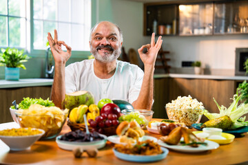 Happy senior Indian foodie enjoying snack-filled table in stylish kitchen setup