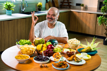 Happy senior Indian foodie enjoying snack-filled table in stylish kitchen setup