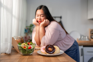 Asian young plus-size woman deciding between green salad and donut. 