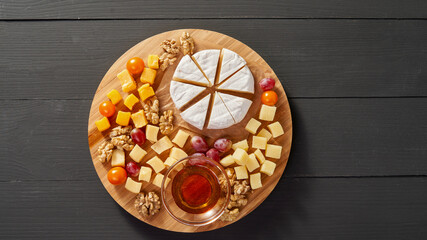 A wooden tray with a variety of cheeses and fruit on it. The tray is placed on a dark wooden table
