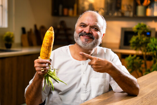 Old Indian foodie relishing roasted corn snack in bright modern kitchen at home