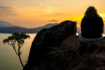 Rear view of woman at sunset on rocks facing Valle de Bravo lake in Mexico from La Pena, golden sky symbolizing serenity, reflection, travel and seasonal identity
