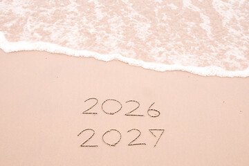 Handwritten inscription 2026 and 2027 with ocean waves on a sandy beach. The concept of Goodbye 2026 and Happy New Year 2027